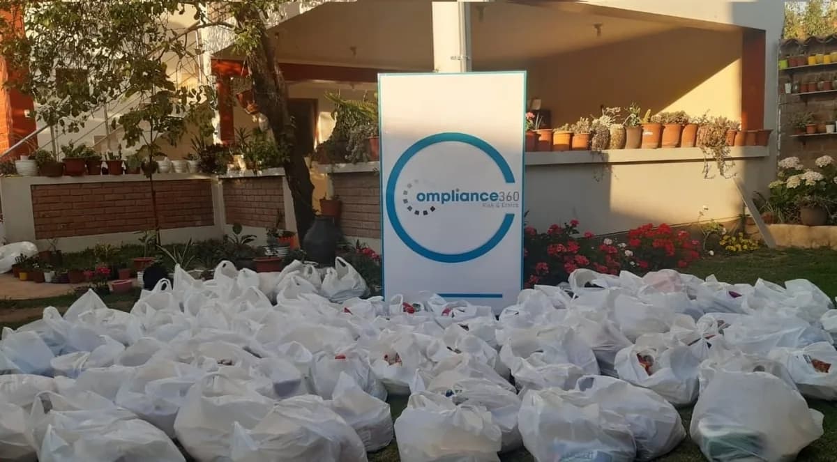 Bolsas de víveres preparadas en un jardín, con el cartel de Compliance360 al fondo